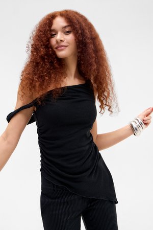 Žena s kučeravými červenými vlasmi, oblečená v čiernom tričku bez ramienok a nohaviciach s prúžkami, usmievajúca sa so striebornými náramkami na zápästí.