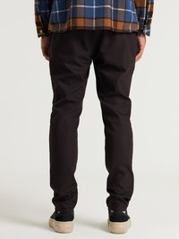 Pantaloni scuri, tagliati su misura, realizzati in tessuto liscio, caratterizzati da una vestibilità slim e una lieve elasticità. Abbinati a sneaker nere e una camicia a quadri.