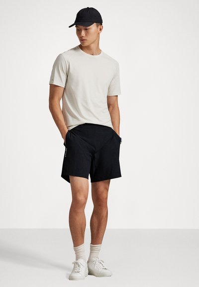 Άνδρας με μαύρο καπέλο, ανοιχτό μπεζ πουκάμισο κοντομάνικο, μαύρες βερμούδες, μπεζ κάλτσες και λευκά αθλητικά παπούτσια, όρθιος με τα χέρια στις τσέπες.