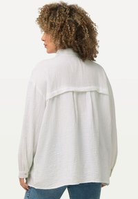 Witte, gestructureerde longsleeve met een rugplooien en knoopdetails op de manchetten, gecombineerd met blauwe jeans.