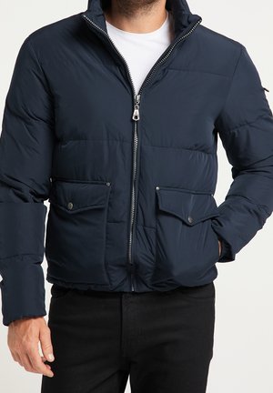 Homme portant une doudoune bleu marine avec fermeture éclair à l'avant et poches à rabat sur une chemise blanche et un pantalon noir.
