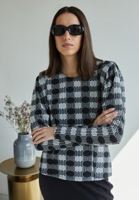 Top de manga longa com padrão em preto e branco e decote redondo; o tecido possui bolinhas em relevo; a modelo usa óculos de sol oversized pretos.