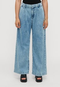 Lyseblå denim vide bukser med høj talje, der har folder, synlige syninger og rå kanter, parret med sorte platform-sandaler.