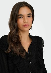 Sort bluse med knaplukning, der har rynkede detaljer langs skuldrene og placket, lavet af tekstureret stof med et prikket mønster.