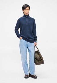 Navy strikket zip-neck sweater, afslappet pasform, lyseblå lige ben jeans, mørke lædersko, holder en olivengrøn taske med brune detaljer.