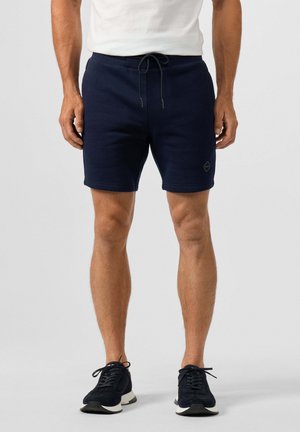 Man draagt marineblauwe shorts met trekkoord, wit t-shirt en zwarte sneakers, staand op een effen grijze achtergrond.