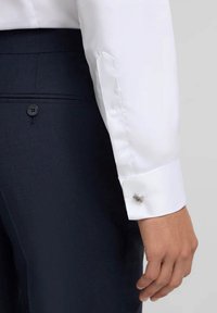Camisa blanca con puños abotonados, que muestra un gemelo de plata. Combinada con pantalones oscuros azul marino, resaltando un ajuste a medida.