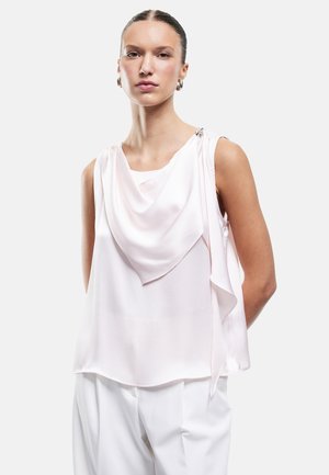 Frau mit zurückgekämmtem Haar, die eine ärmellose, hellrosa drapierte Bluse und weiße Hose vor weißem Hintergrund trägt.