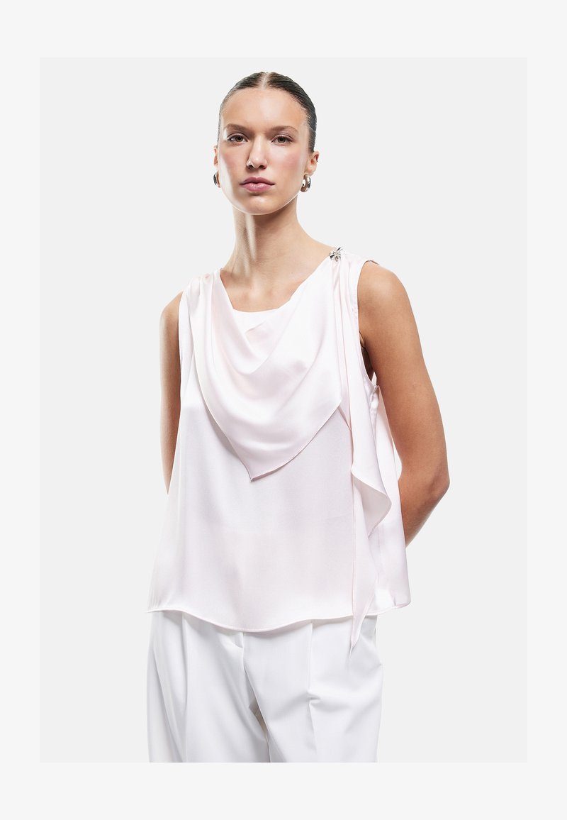 Woman with slicked-back hair wearing sleeveless pale pink draped blouse and white pants against plain white background.