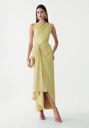 Mujer con vestido drapeado sin mangas de color verde pálido con dobladillo asimétrico, pendientes de oro, pulsera, bolso de mano y tacones con tiras, de pie sobre fondo blanco.