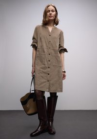 Vestito camicia marrone con maniche arrotolate, due tasche frontali, abbinato a stivali bordeaux al ginocchio e una borsa a tracolla nera e beige.