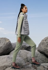 Gilet grigio puffoso, maglione a maniche lunghe verde chiaro e pantaloni verdi. Le calzature combinano tonalità di rosso e nero. Il modello è in piedi su una superficie rocciosa.