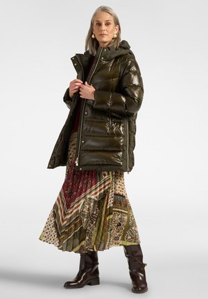 Mujer con un abrigo acolchado verde oscuro brillante sobre una falda con patrones multicolores y botas marrón oscuro, de pie frente a un fondo neutro.