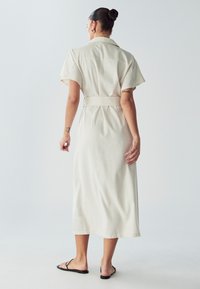 Helles Beige, knielanges Kleid mit kurzen Ärmeln und tailliertem Gürtel. Verfügt über einen klassischen Kragen und eine glatte Textur. Gestylt mit schwarzen Sandalen.