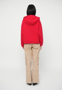 Άτομο με ίσια μαύρα μαλλιά, ντυμένο με κόκκινη φόρμα και μπεζ cargo παντελόνι, που στέκεται με την πλάτη γυρισμένη σε ένα απλό ανοιχτό φόντο.