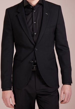Zwarte op maat gemaakte blazer met notch-revers, enkele knoopsluiting, gestructureerde stof en een borstzak aan de linkerkant, gedragen over een zwart shirt.