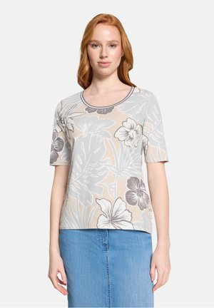 Rødhåret kvinde iført en beige og grå blomstret kortærmet top og en blå denim nederdel, stående foran en ensfarvet baggrund.