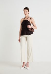 Črn nabrani halter top z oprijetim pasom, v kombinaciji z belimi širokimi hlačami in belimi čevlji s koničastim prstom, drži rjav nahrbtnik.