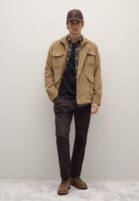 Giacca di velluto a coste marrone chiaro con due tasche frontali sopra una camicia a quadri, t-shirt scura girocollo, pantaloni marroni e stivali marroni.