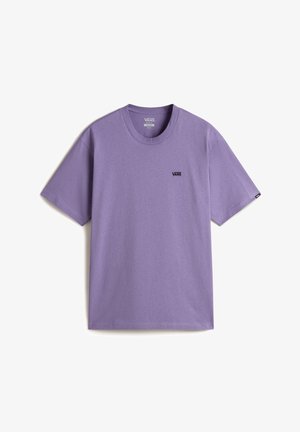 Man draagt een lichtpaars Vans t-shirt met korte mouwen en zwarte gewassen jeans, staat met één hand in de zak.