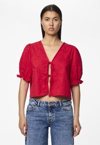Blusa rossa corta con maniche a sbuffo, caratterizzata da un scollo a V e chiusura a fiocco sul davanti, realizzata in tessuto texturizzato. Abbinata a jeans a vita alta.