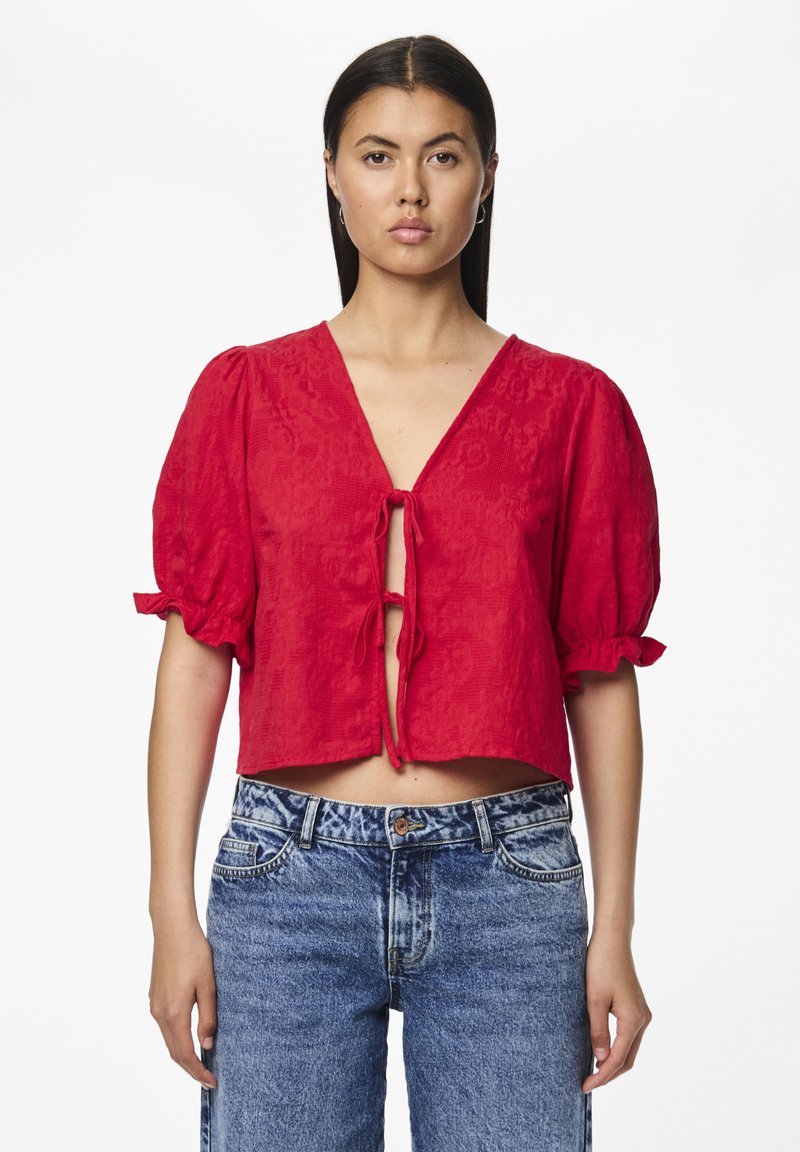 Blusa rossa corta con maniche a sbuffo, caratterizzata da un scollo a V e chiusura a fiocco sul davanti, realizzata in tessuto texturizzato. Abbinata a jeans a vita alta.