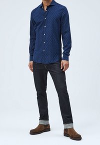 Tummansininen nappipaita, jossa on pieni valkoinen kuvio, pitkät hihat ja kaartuva helma, yhdistetty tummiin farkkuihin ja ruskeisiin mokkanahkaisiin nilkkureihin.