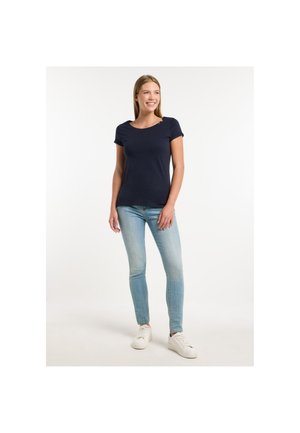 Joven mujer de pie sonriendo, vistiendo una camiseta de manga corta azul marino, vaqueros azul claro y zapatillas blancas sobre un fondo blanco.