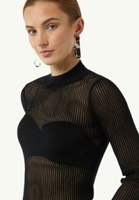 Czarny sweter o prążkowanej fakturze z długimi rękawami i wysokim dekoltem, z przezroczystymi panelami na solidnej czarnej warstwie. Srebrne kolczyki w kształcie kół.