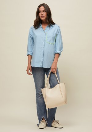 Nainen, jolla on pitkät tummat hiukset, hänellä on sininen paita, etuosastaan halkiot farkuissa, beiget kengät ja hän pitää isoa beigeä käsilaukkua yksiväristä taustaa vasten.