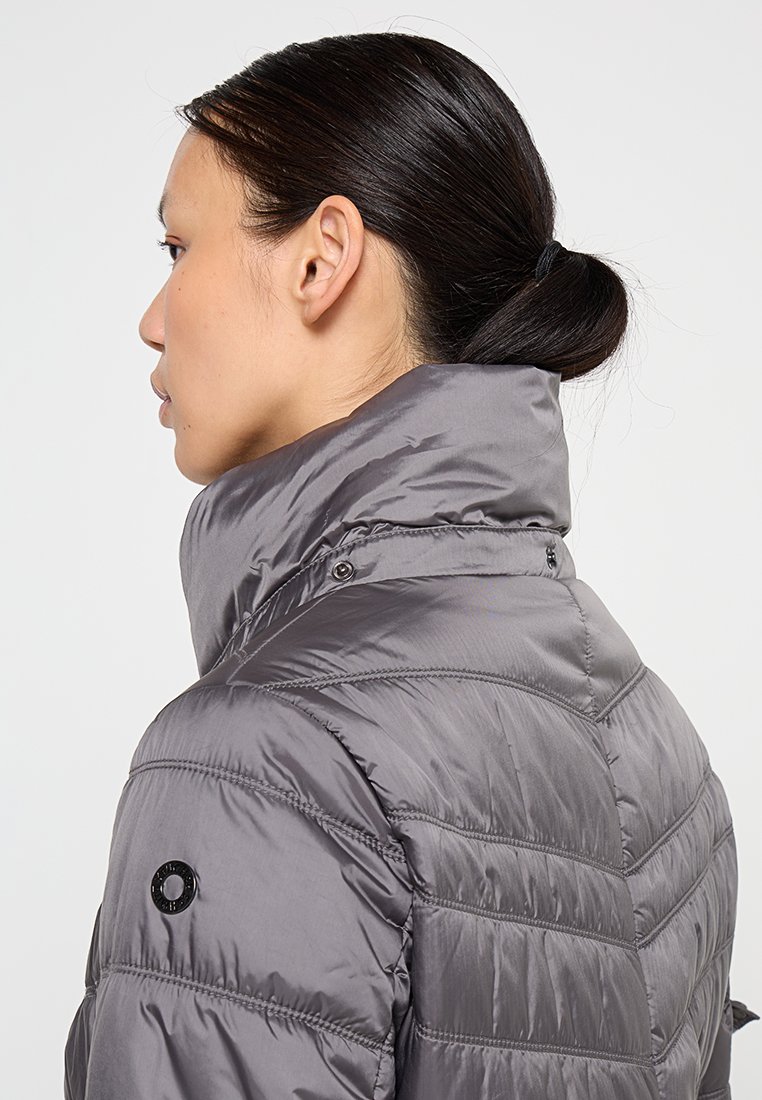 Kobieta o ciemnych włosach związanych w niski kok, ubrana w błyszczącą szaro puchową kurtkę z podniesionym kołnierzem, pokazana z boku i z tyłu na białym tle.