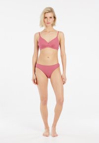 Donna in piedi a piedi nudi che indossa un bikini rosa a coste abbinato, di fronte a uno sfondo bianco uniforme, con una postura rilassata.