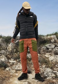 Giacca nera gonfia, pantaloni arancioni con tasche verdi, berretto beige e scarpe nere. Ambientazione all'aperto con terreno roccioso e cielo azzurro.