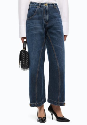 Vaqueros azul oscuro de pierna ancha con detalle de costura frontal, dobladillos con puños y botones laterales, combinados con tacones altos negros puntiagudos y una camisa blanca.