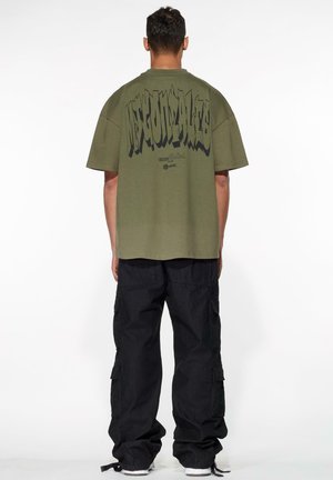 Oliven grønn oversized t-skjorte med stor svart tekst på baksiden. Matchet med svarte cargo-bukser med sidelommer. Uformell passform.