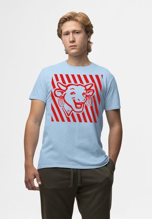 Lyseblå bomulds t-shirt med en rød grafik af en smilende ko med øremærker, sat mod en diagonal rød og hvid stribet baggrund.