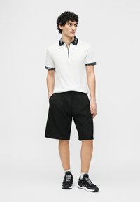 Νεαρός με κατσαρά μαλλιά που φοράει λευκό πόλο με κοντά μανίκια, μαύρη σορτς, μαύρα αθλητικά παπούτσια, στέκεται με τα χέρια στις τσέπες.