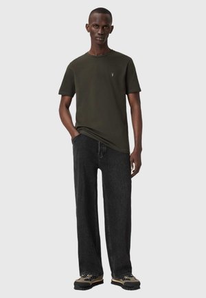 Dunkelgrünes T-Shirt mit kurzen Ärmeln, rundem Ausschnitt und kleinem Logo. Kombiniert mit lässigen schwarzen Jeans und strukturierten Schuhen.