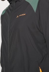 Černá lehká bunda s zelenými ramenními akcenty, boční kapsa na zip a oranžovým logem. Hladký povrch s úzkým střihem.