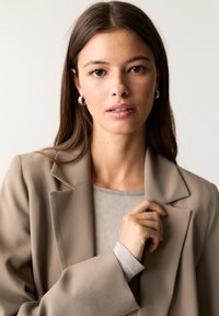 Blazer beige avec une finition texturée, haut gris en dessous et boucles d'oreilles en argent. Le modèle touche le col du blazer.