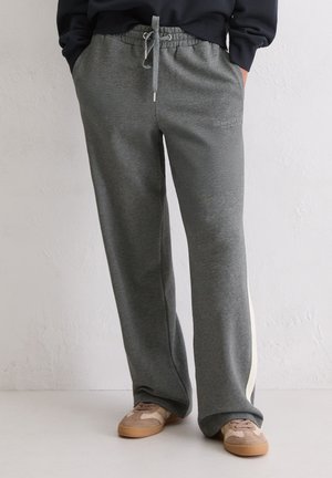 Osoba w szarych spodniach dresowych ze ściągaczem, białym paskiem po boku i beżowych trampkach, stoi przy jednolitej ścianie.