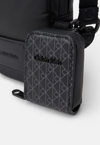 Geantă neagră Calvin Klein cu un portofel texturat, cu un model gri atașat, având un fermoar și o marcă subtilă pe ambele articole.