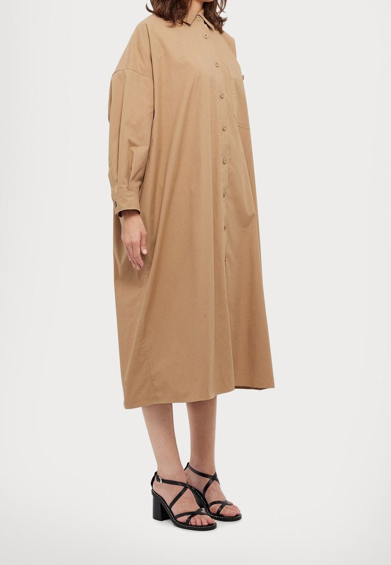 Vestido caqui de botones con mangas largas, corte suelto y bolsillo en el pecho izquierdo. Combinado con sandalias negras de tacón alto con tiras.
