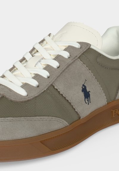 Καθημερινό sneaker με πάνελ από βελούδινο ύφασμα σε ελαιώνιο πράσινο και μπεζ, λευκά κορδόνια, καφέ λαστιχένια σόλα και σκούρο λογότυπο παίκτη πόλο στο πλάι.