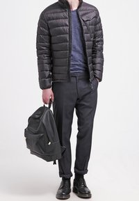 Chaqueta acolchada negra con diseño acolchado horizontal, camisa azul marino con rayas, pantalones oscuros a medida y mochila negra con textura.