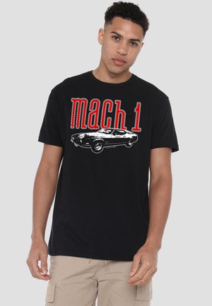 Young man wearing black "mach 1" t-shirt with vintage car graphic, standing against a plain light gray background.