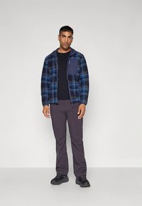 Modra in črno karo jakna iz flisa z zadrgo spredaj in žepom na prsih, kombinirana s temno sivimi hlačami in črnimi supergami.