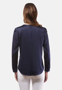 Blouse en satin bleu marine à manches longues avec encolure ronde, fermeture par bouton au dos et léger froncé aux épaules ; texture lisse et ourlet arrondi.