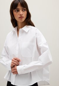 Femme aux longs cheveux bruns portant une chemise blanche boutonnée ample, regardant sur le côté devant un fond clair.