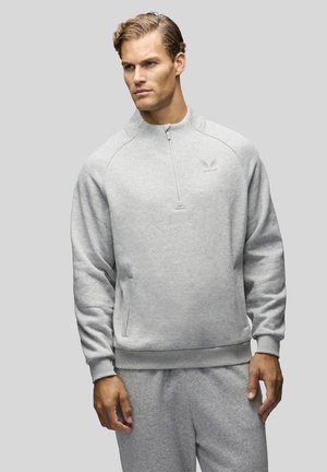 Homme portant un sweat-shirt gris clair à fermeture éclair quart-zip et un pantalon de survêtement assorti, debout devant un fond gris uni.